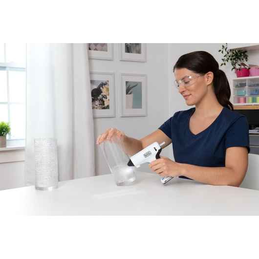 BLACK+DECKER BCGL115 hot glue gun in use on a glass vase, indoor craft setting, side angle.