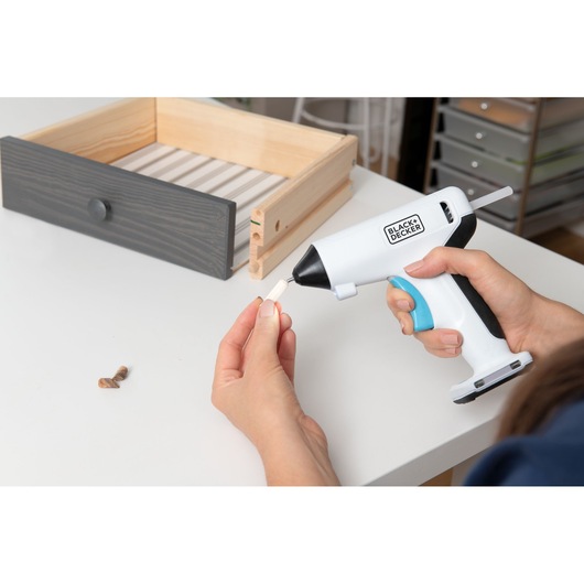 BLACKDECKER BCGL115 cordless glue gun held by hand, applying glue to wooden piece near drawer on tabletop.