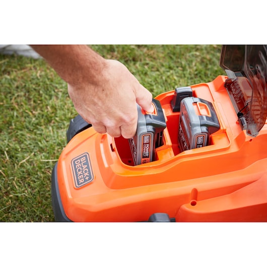 Photo of BLACK+DECKER BCMW3318L2 orange lawn mower with hand inserting battery, close-up outdoors.