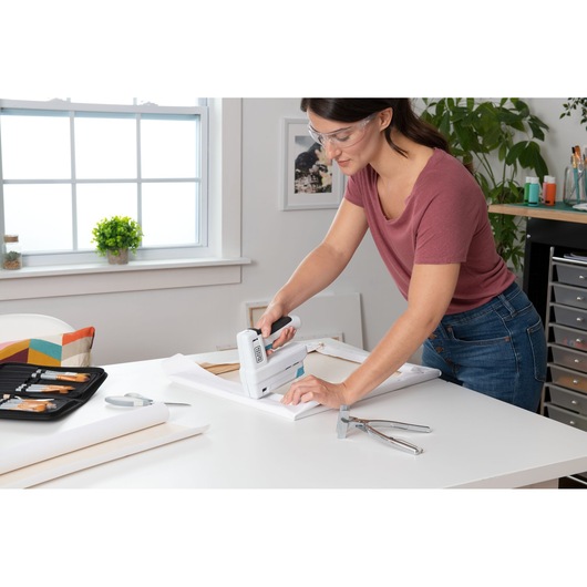 Photo of person using BLACK+DECKER BCN115 staple gun on a canvas frame in a bright home workspace.