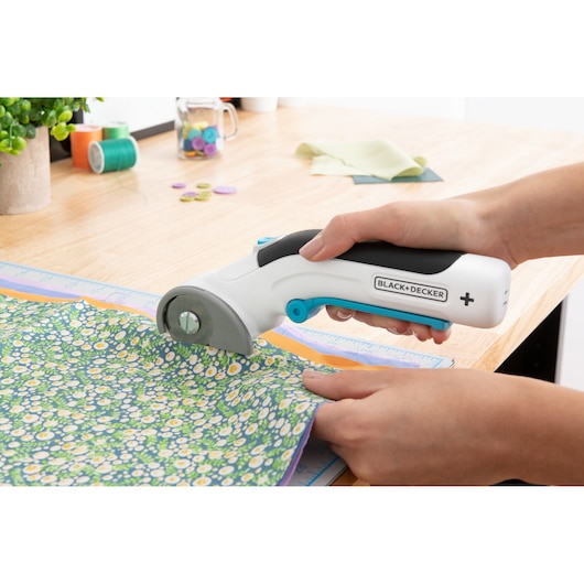 BLACK+DECKER BCRC115 rotary fabric cutter trimming floral fabric on a wooden table, hand-held operation.