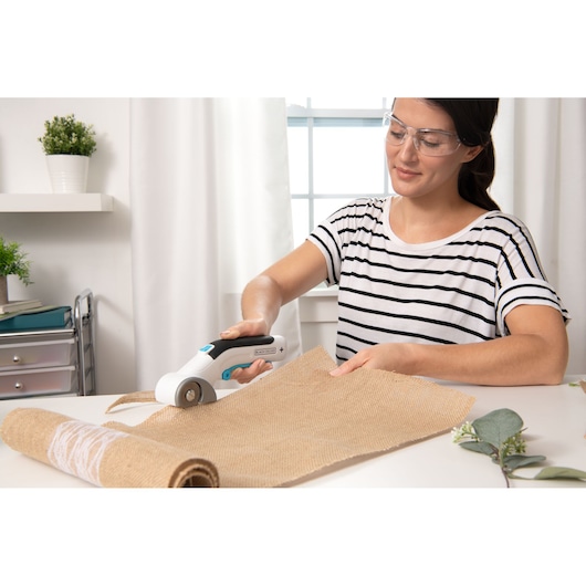 Photo of BLACK+DECKER BCRC115 rotary cutter being used to cut burlap fabric on a white table indoors.