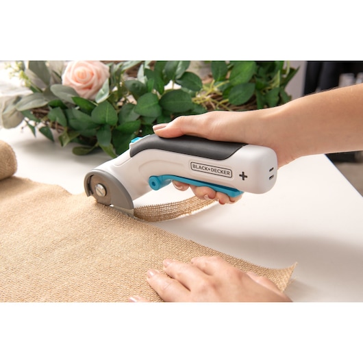Photo of BLACK+DECKER BCRC115 cordless fabric cutter held in hand, cutting burlap cloth on a table.
