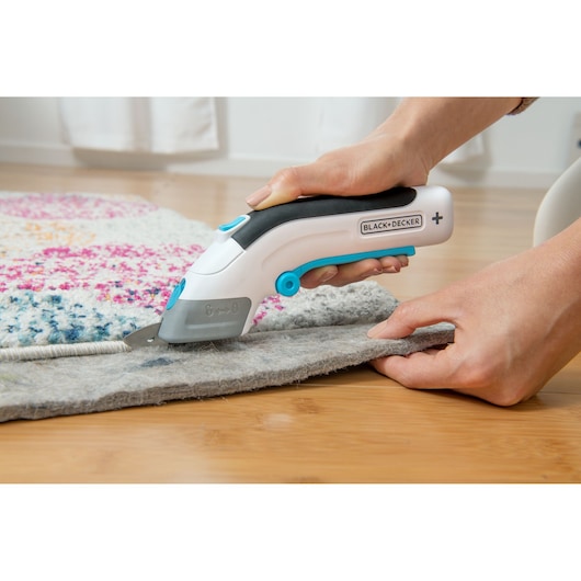 BLACK+DECKER BCSC115 electric cutter trimming gray carpet on wooden floor, close-up side view.