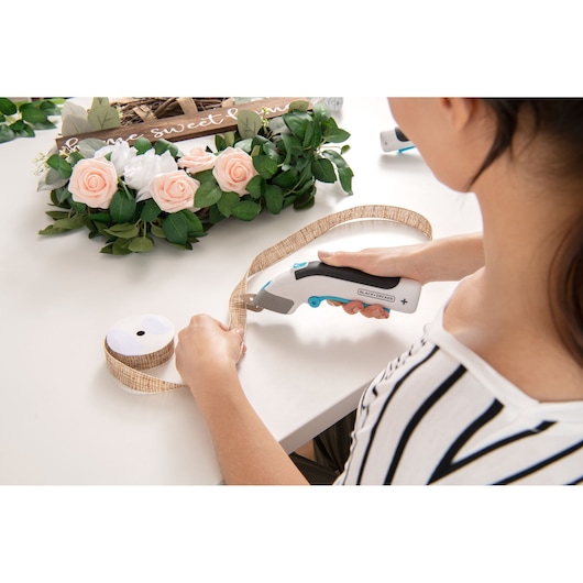 Photo of person using BLACK+DECKER BCSC115 electric scissors to cut decorative ribbon, flowers and wreath on table.