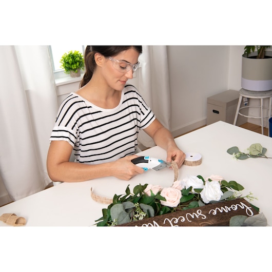 Photo of person using BLACKDECKER BCSC115 cordless craft scissors to cut ribbon for floral DIY project on white table.