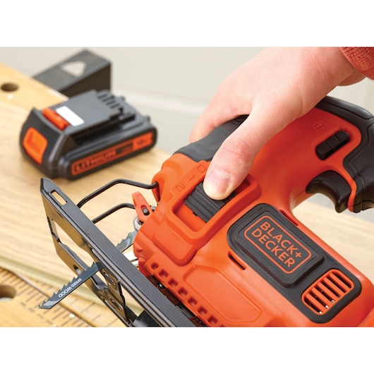 Photo of BLACK+DECKER BDCJS18 cordless jigsaw in use on wood, showing blade, handle, and visible battery charger.