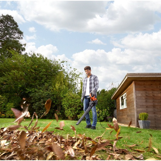 BLACKDECKER BEBLV301 leaf blower in use outdoors, blowing leaves near a wooden shed on a grassy lawn.