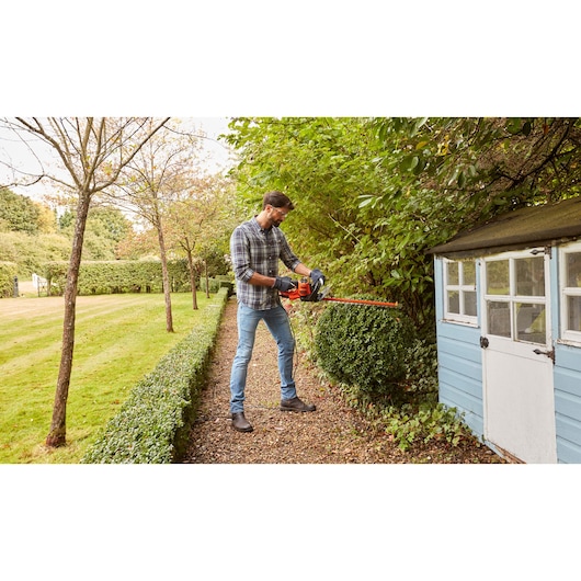 Photo of a person using BLACKDECKER BEHTS401 hedge trimmer to cut hedges beside a small garden shed.