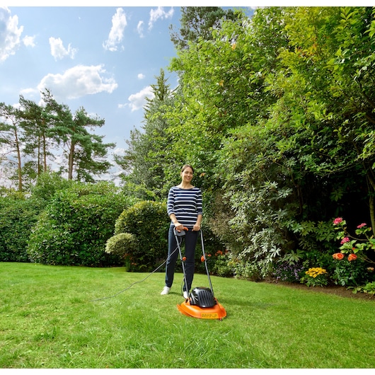Photo of person using BLACK+DECKER BEMWH551 orange electric lawn mower in garden surrounded by green shrubs and flowers.