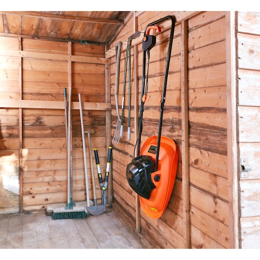 Photo of BLACK+DECKER BEMWH551 orange and black hover mower hanging on a wooden shed wall with garden tools nearby.