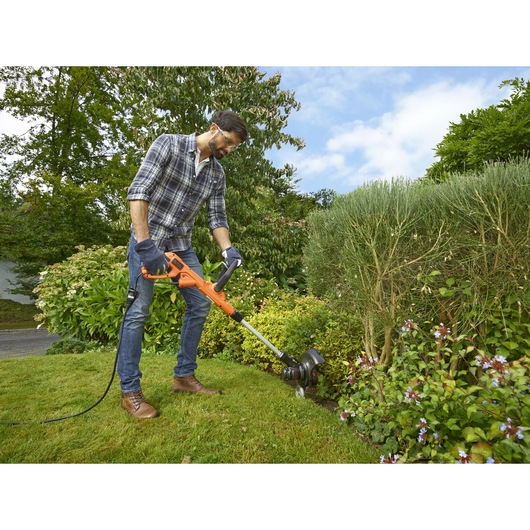 Person using BLACKDECKER BESTE628 electric grass trimmer in a garden, trimming lawn edges near bushes.