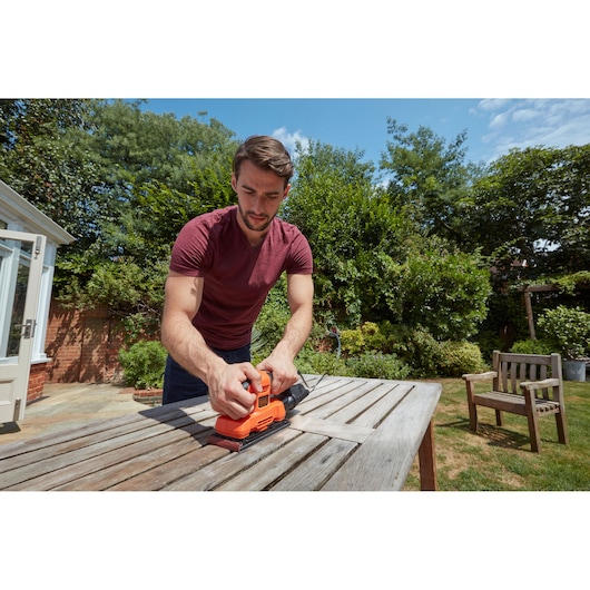 Person using BLACKDECKER BEW220 electric sander on outdoor wooden table in garden.
