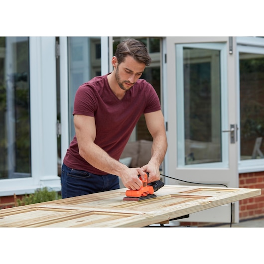 Person using BLACKDECKER BEW220 orange electric sander on wooden door outdoors.