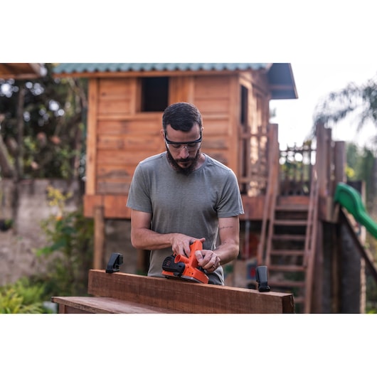 Person using BLACK+DECKER BEW712 electric sander on wooden board outdoors, house and slide in background.