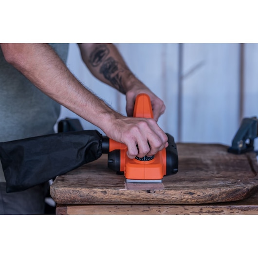 Photo of BLACKDECKER BEW712 hand planer in use on wood, showing hands operating the orange tool with dust bag attached.