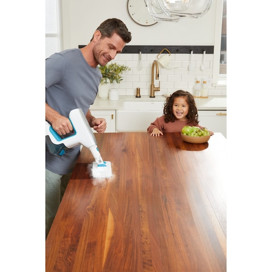BLACKDECKER BHSM15FX08 steam cleaner used on wooden kitchen table, fruit bowl visible in background.