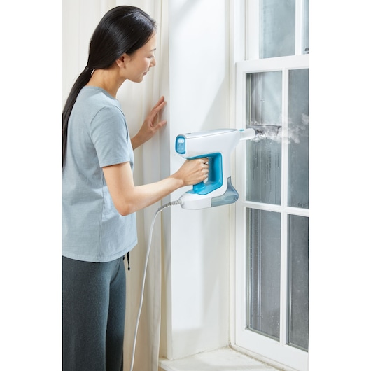 BLACKDECKER BHSM15FX08 steam cleaner held to clean window glass, featuring blue and white handheld design.