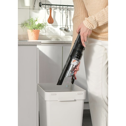 A person is emptying the dust container of a BLACK+DECKER handheld vacuum cleaner into a white trash bin in a kitchen setting.