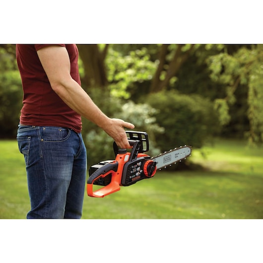 Photo of person holding BLACKDECKER GKC3630L20 cordless chainsaw, orange and black, outdoor garden setting.