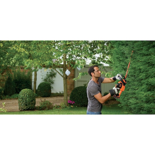 A person using a BLACK+DECKER cordless hedge trimmer to prune a large green hedge in a garden, wearing gloves and a grey shirt. The person's face is blurred.