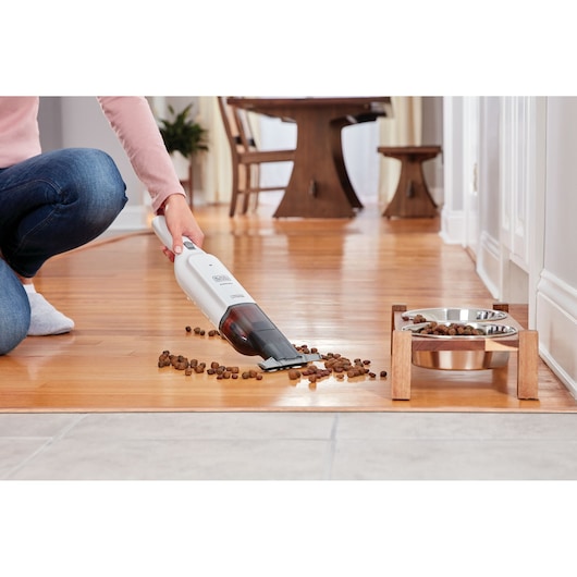BLACK+DECKER HLVC315B11 handheld vacuum cleaning pet food from wooden floor near pet bowls, side angle.