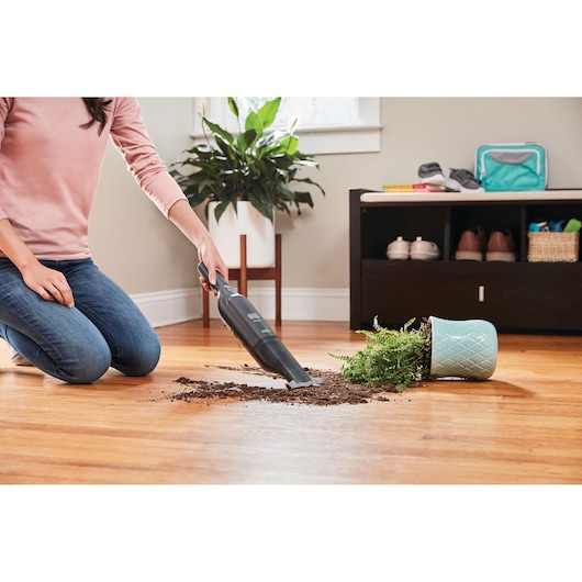 BLACK+DECKER HLVC320B11 handheld vacuum cleaning spilled soil from fallen plant on hardwood floor.