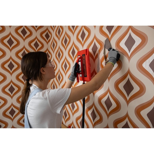A woman removes wallpaper using the BLACK+DECKER® 2400W Wallpaper Stripper.