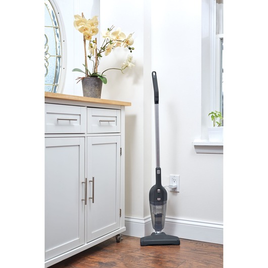 BLACKDECKER NSVA315J cordless stick vacuum cleaner upright against a white wall near a cabinet, indoor setting.