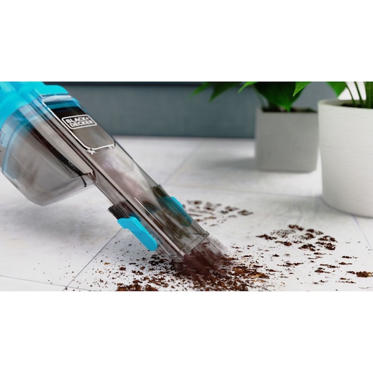 BLACK+DECKER NVD215J handheld vacuum cleans dirt on white floor near potted plants, angled side view.