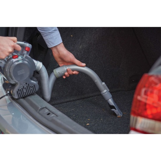 A person is using a Black+Decker handheld vacuum cleaner with a flexible hose attachment to clean the interior of a car trunk.
