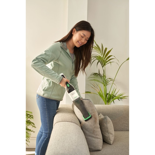Photo of person using BLACKDECKER REVHV8C handheld vacuum to clean a light gray sofa in a modern living room.