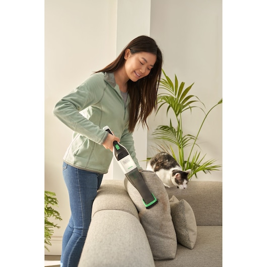 Photo of person using BLACKDECKER REVHV8C handheld vacuum to clean a sofa near a cat in a living room.