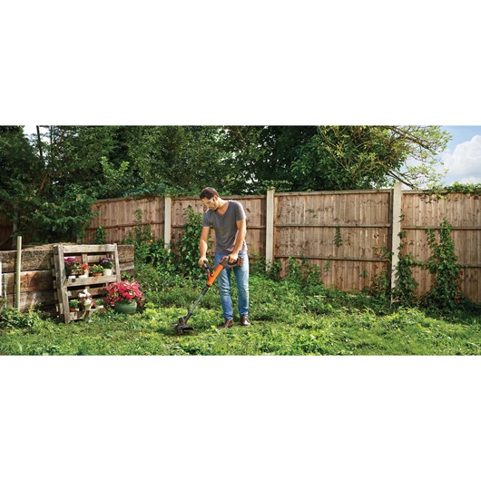 A person using a BLACK+DECKER cordless grass trimmer in a garden, surrounded by a wooden fence and greenery. There are flowers planted in containers on the left side.