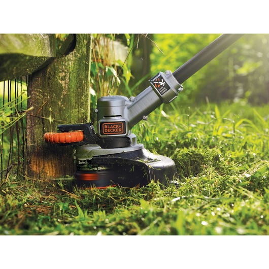 A BLACK+DECKER grass trimmer being used to cut grass and trim edges near a wooden fence in a garden.